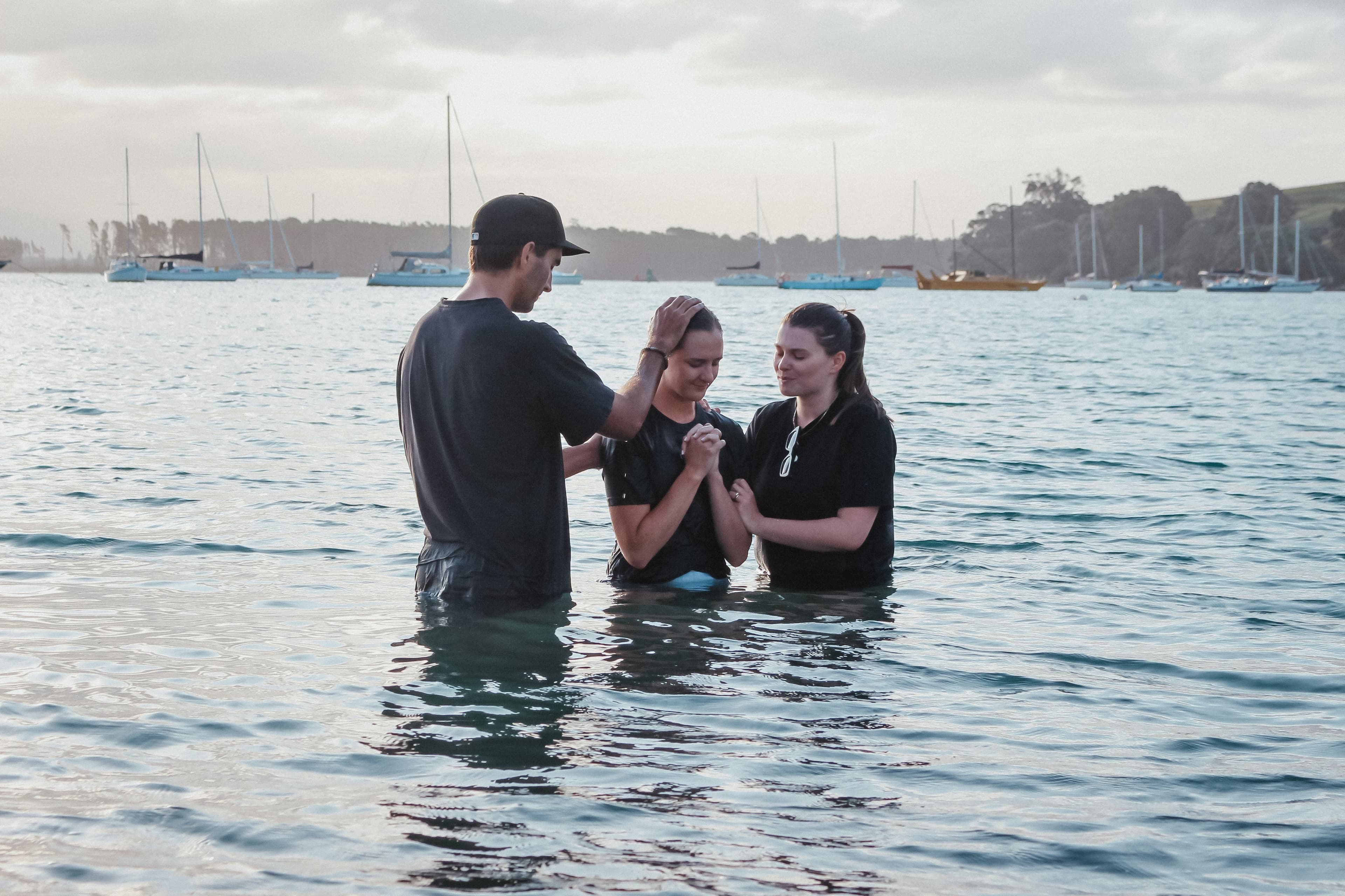 Man and woman laying hands in prayer on the shoulders of a young woman who has just been baptised at Pilot Bay in Tauranga with moored boats in the background